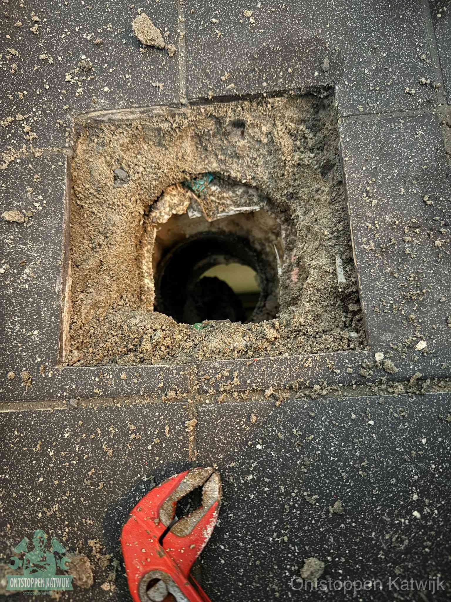 Een geopend rioolputje in een betontegelvloer met losgemaakt rooster en een zichtbare ronde afvoerbuis, het achterhalen waar de afvoerverstopping zit in Katwijk