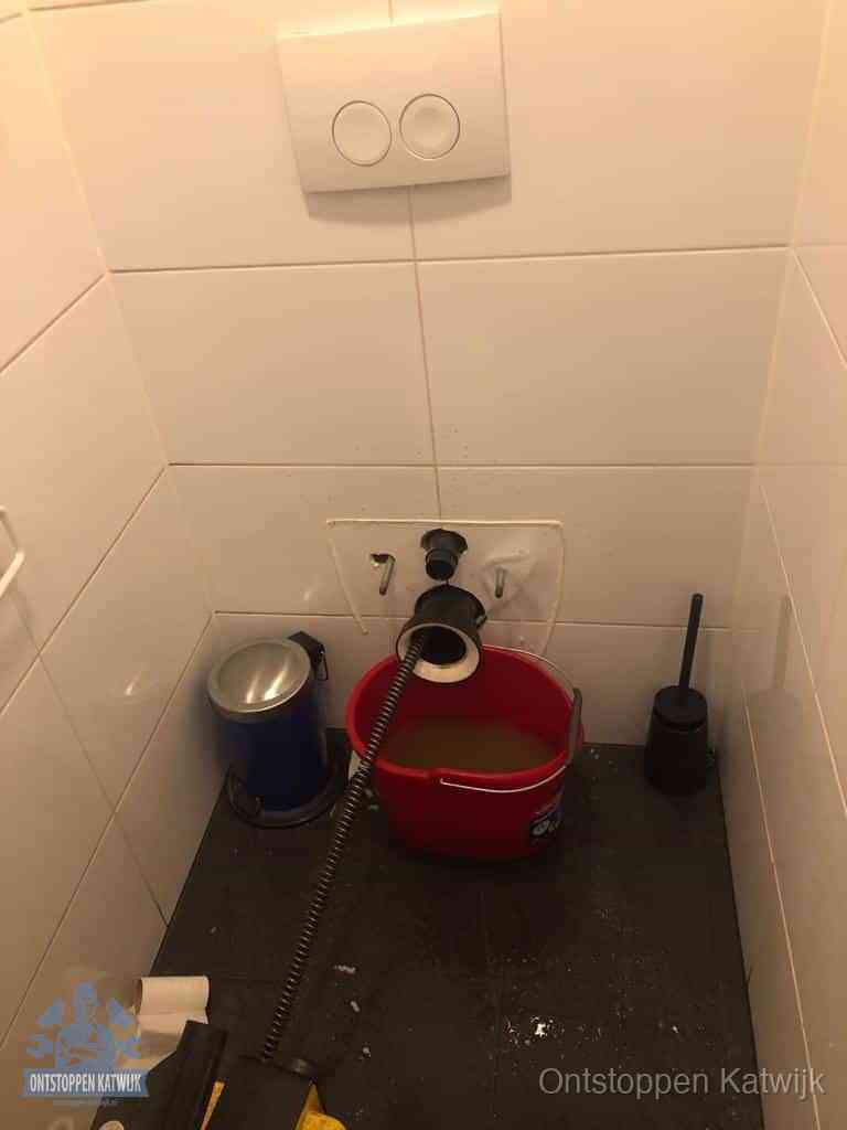 Een ontstopte wand-wc met verwijderde toiletpot waar een ontstoppingsveer in de wanduitgang verdwijnt, het achterhalen waar de verstopping zit in Katwijk