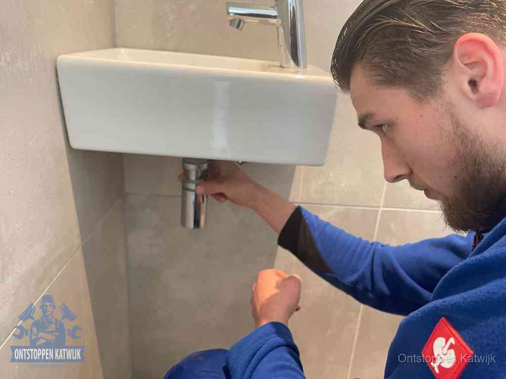 Een monteur draait met de hand de zwanenhalssifon onder een fonteinwasbak in de toiletruimte los om de bron van rioollucht in Katwijk te vinden