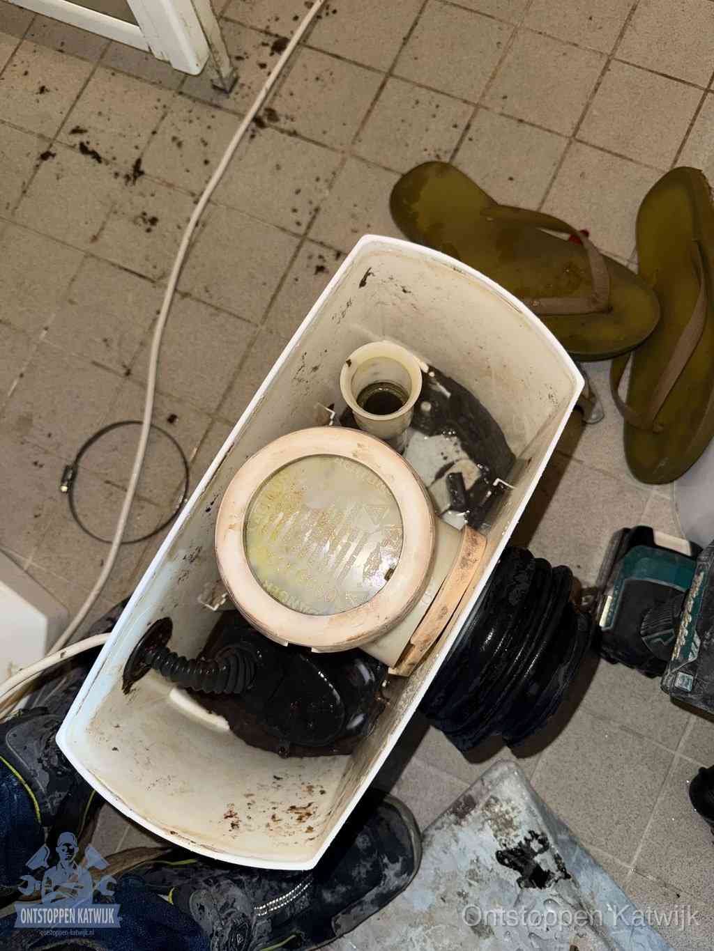 Een gedemonteerde toiletpot op de vloer met ontstoppingsveer en gereedschap ernaast, het verwijderen van een vastzittend voorwerp uit de wc in Katwijk