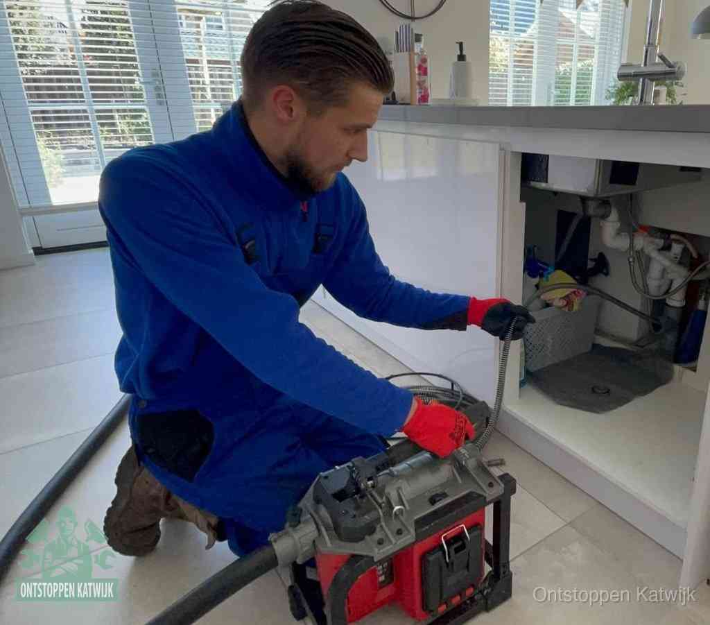 Een monteur met rode handschoenen knielt in een keuken en voert een elektrische ontstoppingsveer in tijdens het professioneel afvoer ontstoppen in Katwijk